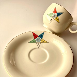 Peut inclure: Une tasse à thé et une soucoupe en céramique blanche avec une bordure dorée. La tasse et la soucoupe présentent une étoile à cinq branches avec un symbole maçonnique au centre.
