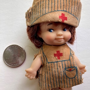 May include: A vintage doll dressed as a nurse with a brown and white striped uniform, a red cross on the chest, and a brown fabric hat with a red cross. The doll has brown hair, blue eyes, and white shoes.