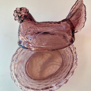 May include: A vintage, amethyst-coloured glass hen-on-nest dish. The hen-shaped lid features detailed feather and comb textures. The nest-shaped base has a woven basket design. This decorative piece is ideal for kitchen or farmhouse decor.