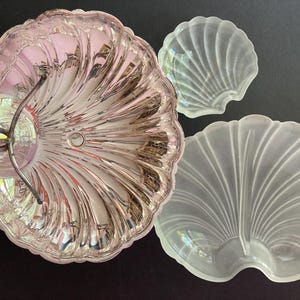 Könnte beinhalten: Ein silberfarbene, muschelförmige Servierschale mit Griff und zwei kleinere, mattierte Glasschalen in Muschelform. Die Schalen sind auf einer dunklen Oberfläche angeordnet. Die Servierschale hat eine reflektierende Oberfläche.