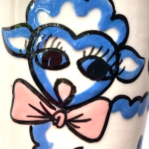 May include: A blue and white ceramic mug with a hand-painted illustration of a sheep with eyelashes and a pink bow. The sheep is wearing a blue bow and has a white face.
