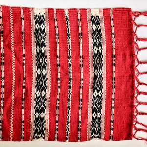 Op de afbeelding: Rood, zwart en wit geweven tapijt met franjes. Het tapijt heeft een geometrisch patroon en is gemaakt van een zacht, getextureerd materiaal.