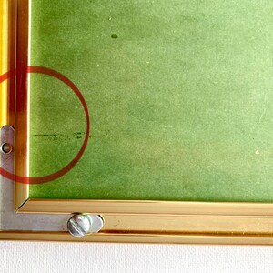 May include: Close-up of a gold picture frame with a green mount. The frame has a visible screw on the bottom right corner.