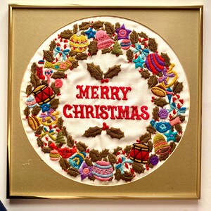 May include: A framed embroidered Christmas wreath with a white background and a gold frame. The wreath is decorated with colourful ornaments, holly leaves, and candy canes. The text "Merry Christmas" is embroidered in red on the wreath.