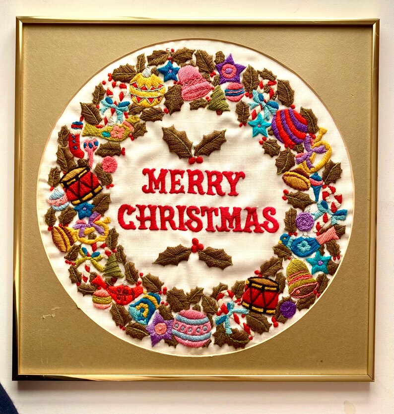 May include: A framed embroidered Christmas wreath with a white background and a gold frame. The wreath is decorated with colourful ornaments, holly leaves, and candy canes. The text "Merry Christmas" is embroidered in red on the wreath.