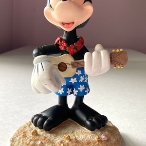 May include: A figurine of Mickey Mouse dressed in Hawaiian attire, playing a ukulele. He is wearing a lei of red flowers and blue shorts with white flowers. He is standing on a sandy base.