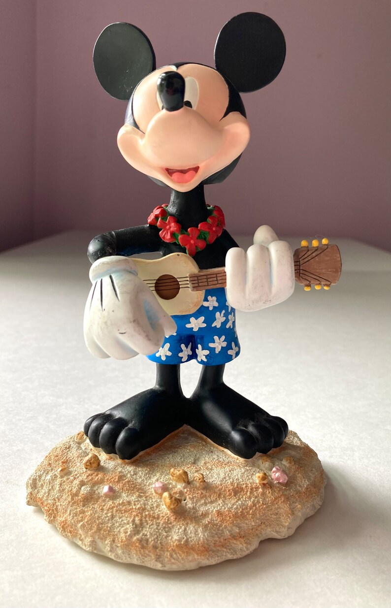 May include: A figurine of Mickey Mouse dressed in Hawaiian attire, playing a ukulele. He is wearing a lei of red flowers and blue shorts with white flowers. He is standing on a sandy base.