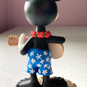 May include: A black and white Mickey Mouse figurine wearing a blue and white floral patterned swimsuit and a red flower lei. He is holding a ukulele and standing on a tan sand base with a pink seashell.
