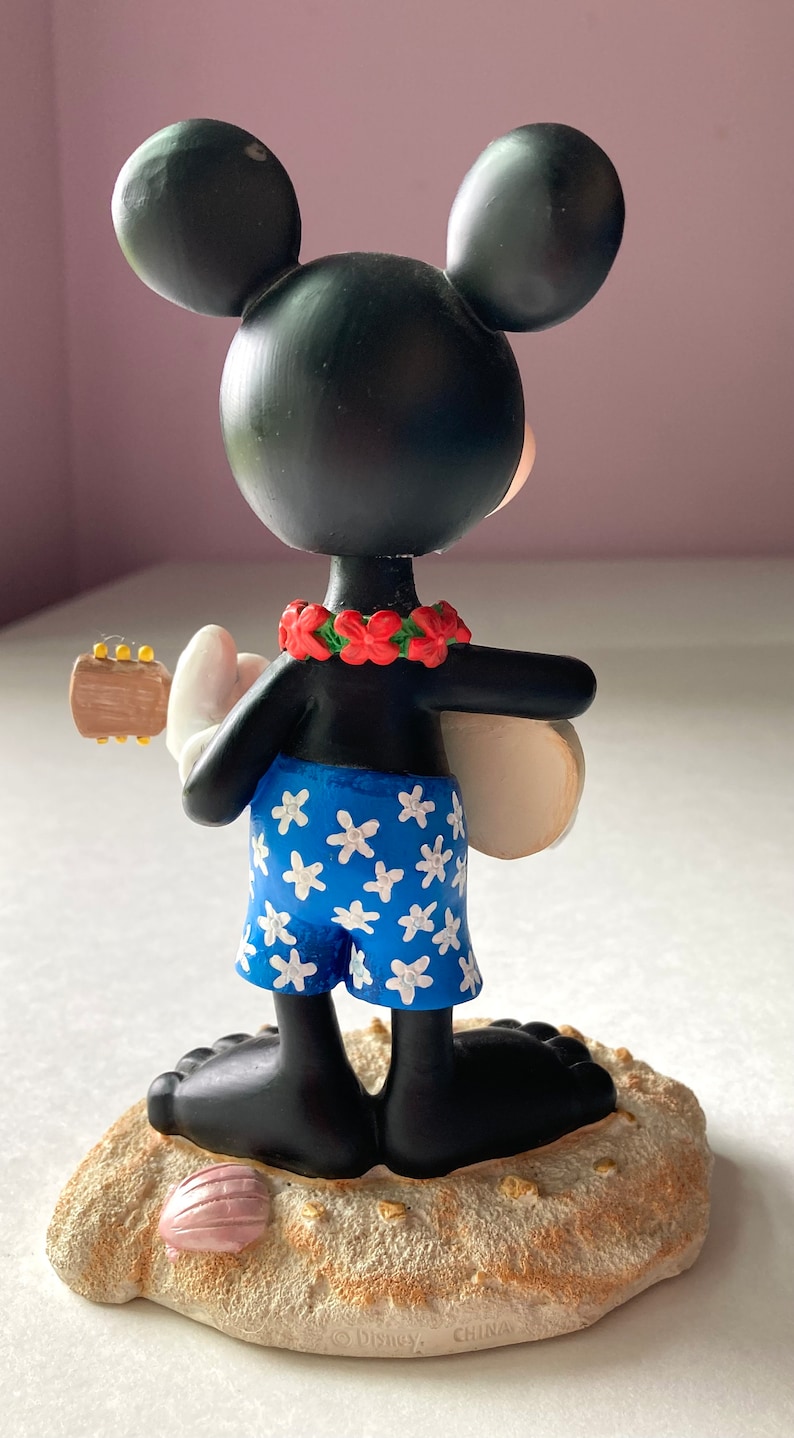 May include: A black and white Mickey Mouse figurine wearing a blue and white floral patterned swimsuit and a red flower lei. He is holding a ukulele and standing on a tan sand base with a pink seashell.