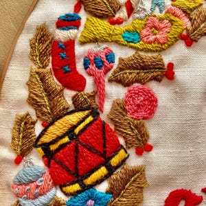 May include: Embroidered Christmas wreath featuring a red and yellow drum, a red stocking, a blue and pink bauble, and green holly leaves. The wreath is on a white background with the word "Christmas" embroidered in red.