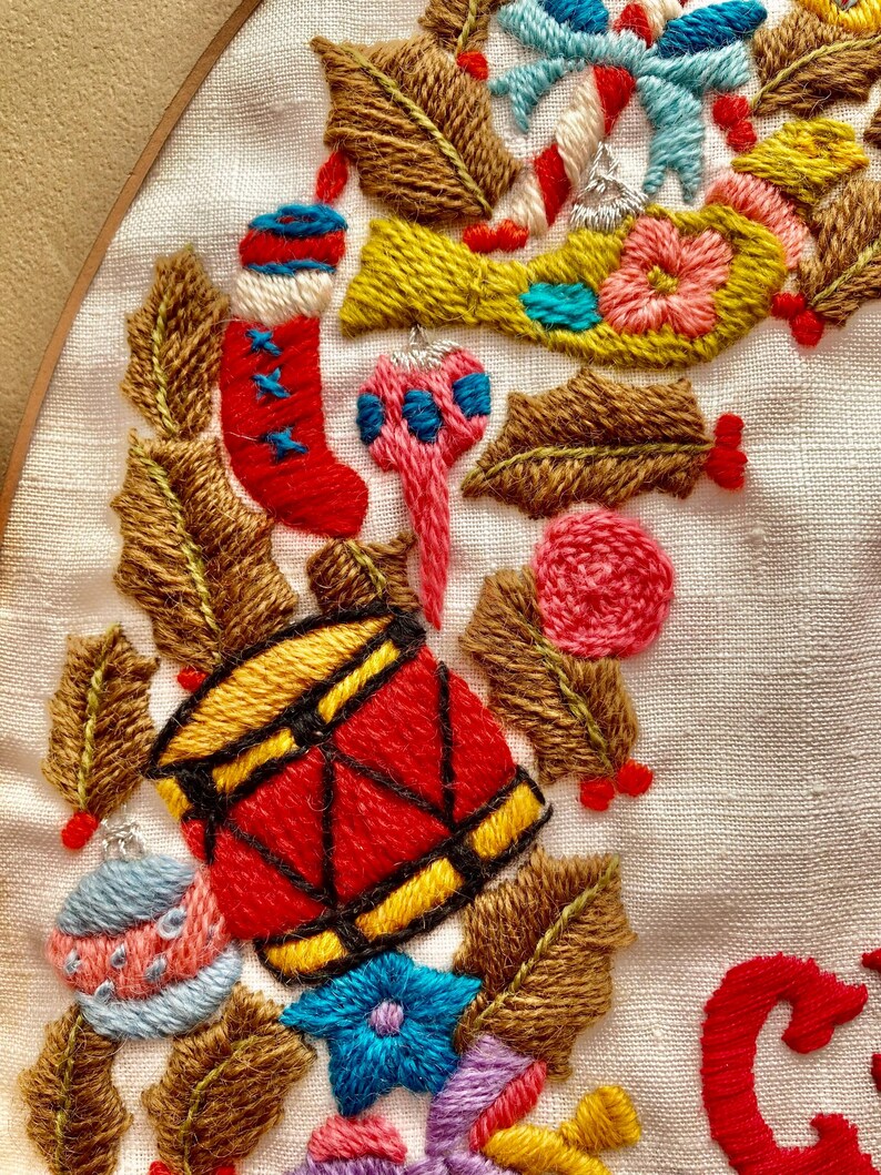 May include: Embroidered Christmas wreath featuring a red and yellow drum, a red stocking, a blue and pink bauble, and green holly leaves. The wreath is on a white background with the word "Christmas" embroidered in red.