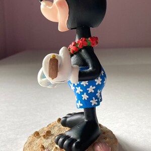 May include: A black and white Mickey Mouse figurine wearing a red lei and blue and white patterned swim trunks. He is holding a white surfboard with a brown fin. The figurine is standing on a tan base with brown and pink seashells.