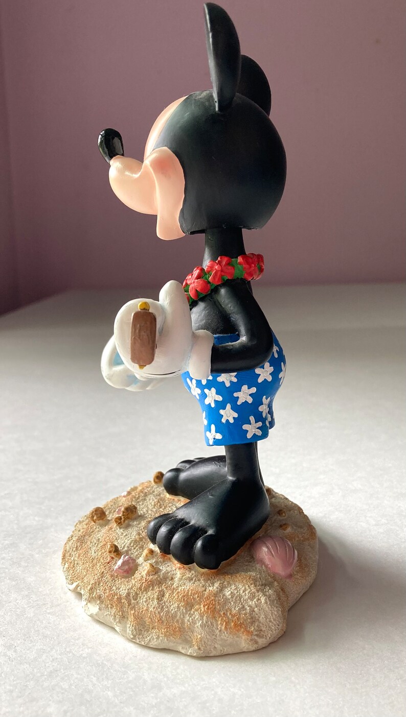 May include: A black and white Mickey Mouse figurine wearing a red lei and blue and white patterned swim trunks. He is holding a white surfboard with a brown fin. The figurine is standing on a tan base with brown and pink seashells.