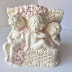 Könnte beinhalten: Ein weißer Keramik-Pflanztopf in Form von drei Cherubinen, die in einem Korb sitzen. Die Cherubinen sind mit rosa und gelben Blumen bemalt.