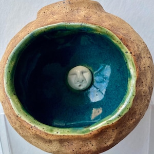Peut inclure: Un vase en céramique fait à la main avec un extérieur brun texturé. L'intérieur présente une glaçure bleu-vert profond avec un bord vert. Une petite sculpture blanche en forme de visage est centrée à l'intérieur.