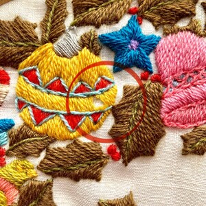 May include: Close-up of a hand-stitched embroidery design featuring a yellow, red and blue Christmas bauble surrounded by brown holly leaves. The bauble has a white top and a red and blue pattern.