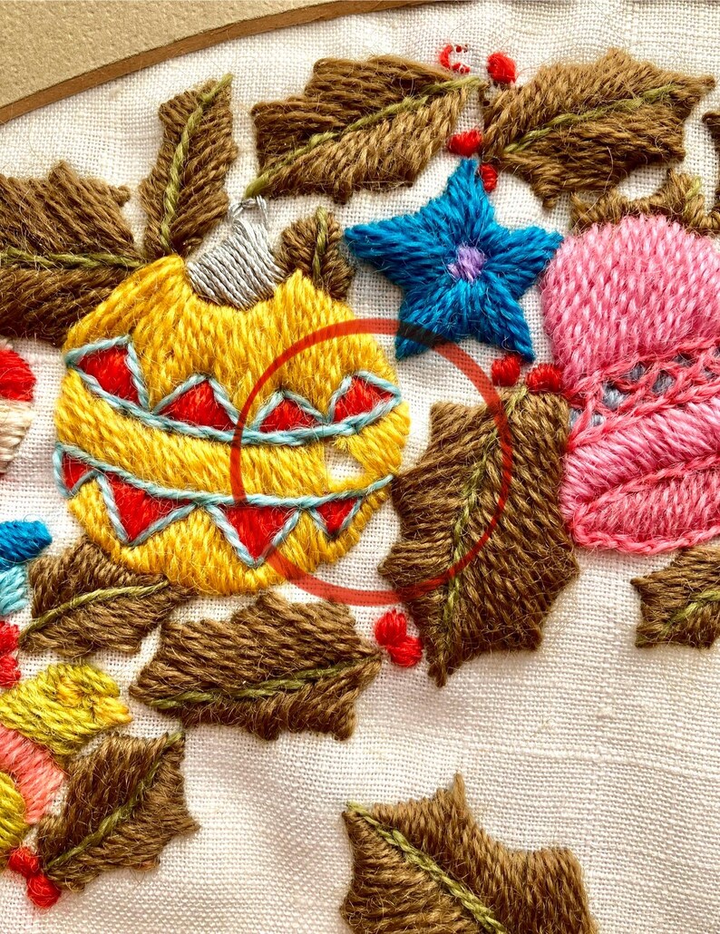 May include: Close-up of a hand-stitched embroidery design featuring a yellow, red and blue Christmas bauble surrounded by brown holly leaves. The bauble has a white top and a red and blue pattern.