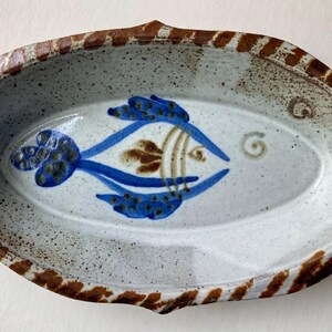 Può includere: Piatto da portata ovale in ceramica con un disegno di pesce dipinto a mano. Il pesce è blu e marrone, su uno sfondo bianco e grigio maculato. Il bordo è marrone con un motivo a strisce. Un pezzo decorativo per servire antipasti o snack.