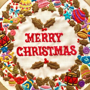 May include: A white fabric Christmas wreath with brown holly leaves, red berries, and colourful ornaments embroidered around the words "Merry Christmas".