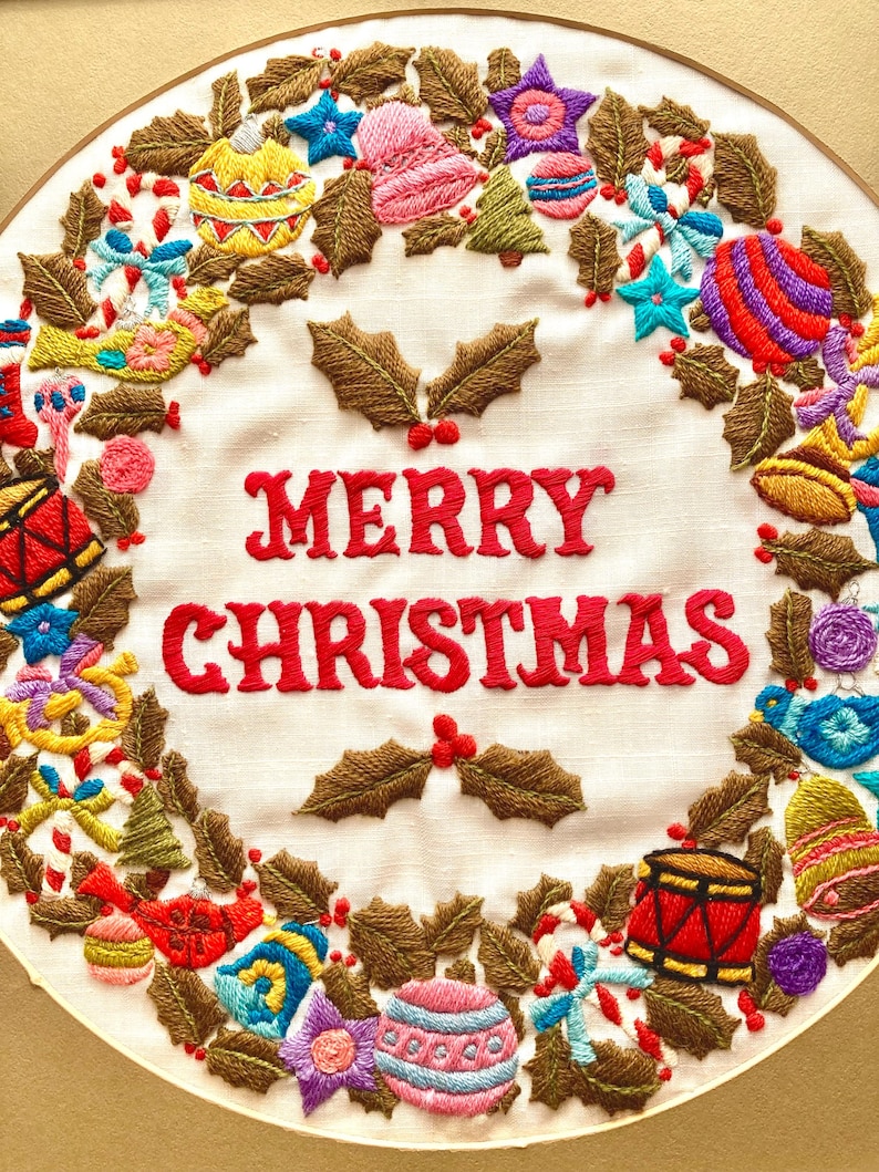 May include: A white fabric Christmas wreath with brown holly leaves, red berries, and colourful ornaments embroidered around the words "Merry Christmas".
