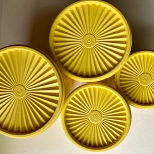 Può includere: Quattro coperchi di plastica gialla di diverse dimensioni sono disposti. Ogni coperchio presenta un design circolare centrale con linee radiali, che formano un motivo a raggiera. I coperchi sono probabilmente per contenitori per alimenti.
