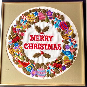 May include: A framed embroidered Christmas wreath with a white background and a red "Merry Christmas" message surrounded by a wreath of green holly leaves and colourful ornaments.