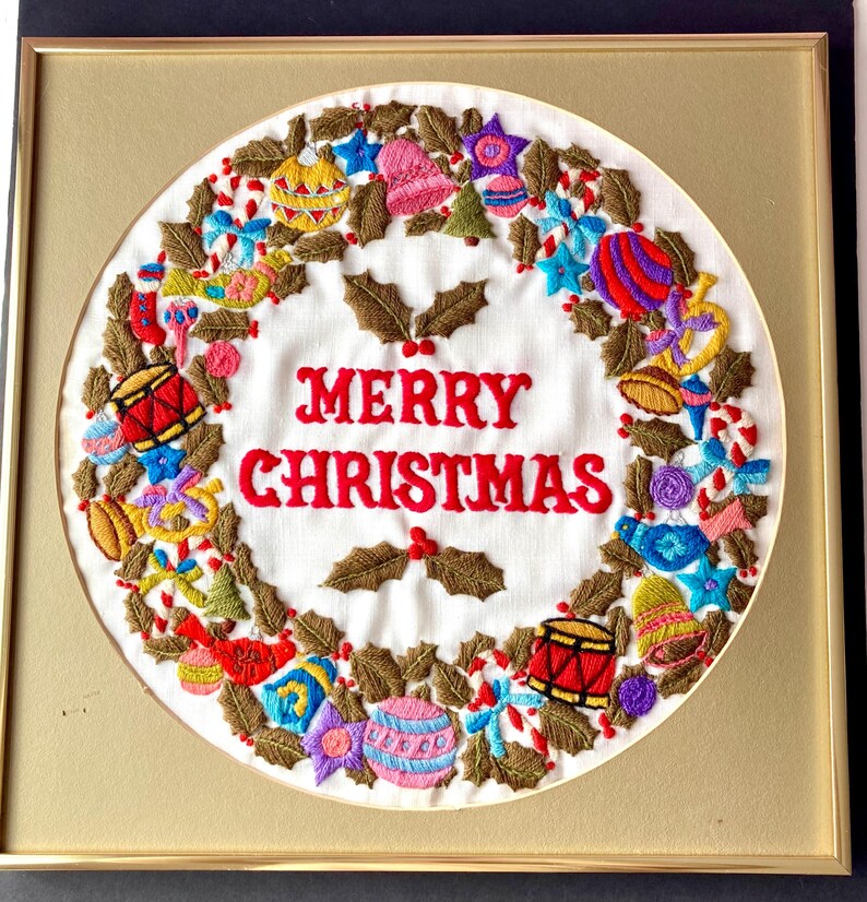May include: A framed embroidered Christmas wreath with a white background and a red "Merry Christmas" message surrounded by a wreath of green holly leaves and colourful ornaments.