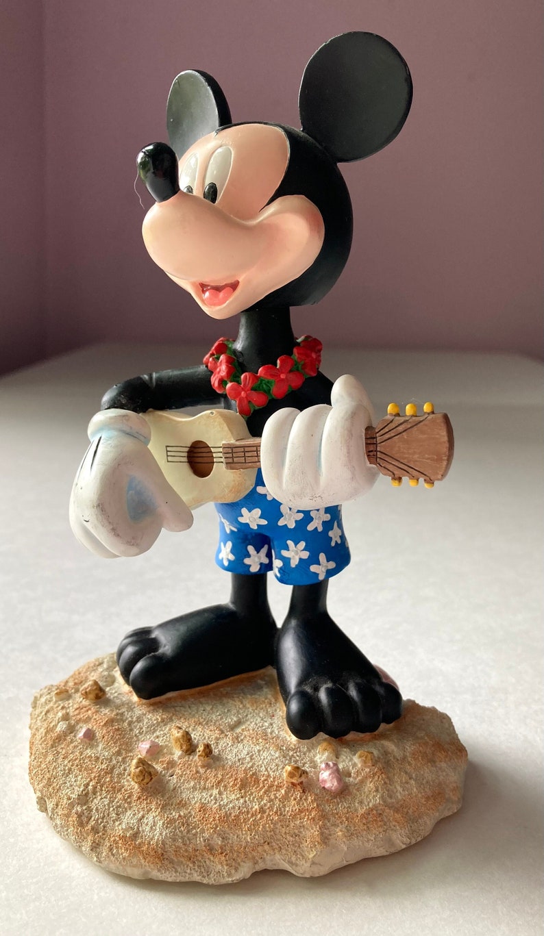 May include: A black and white Mickey Mouse figurine wearing a red flower lei and blue and white floral swim trunks. He is holding a brown ukulele and standing on a tan and pink sand base.
