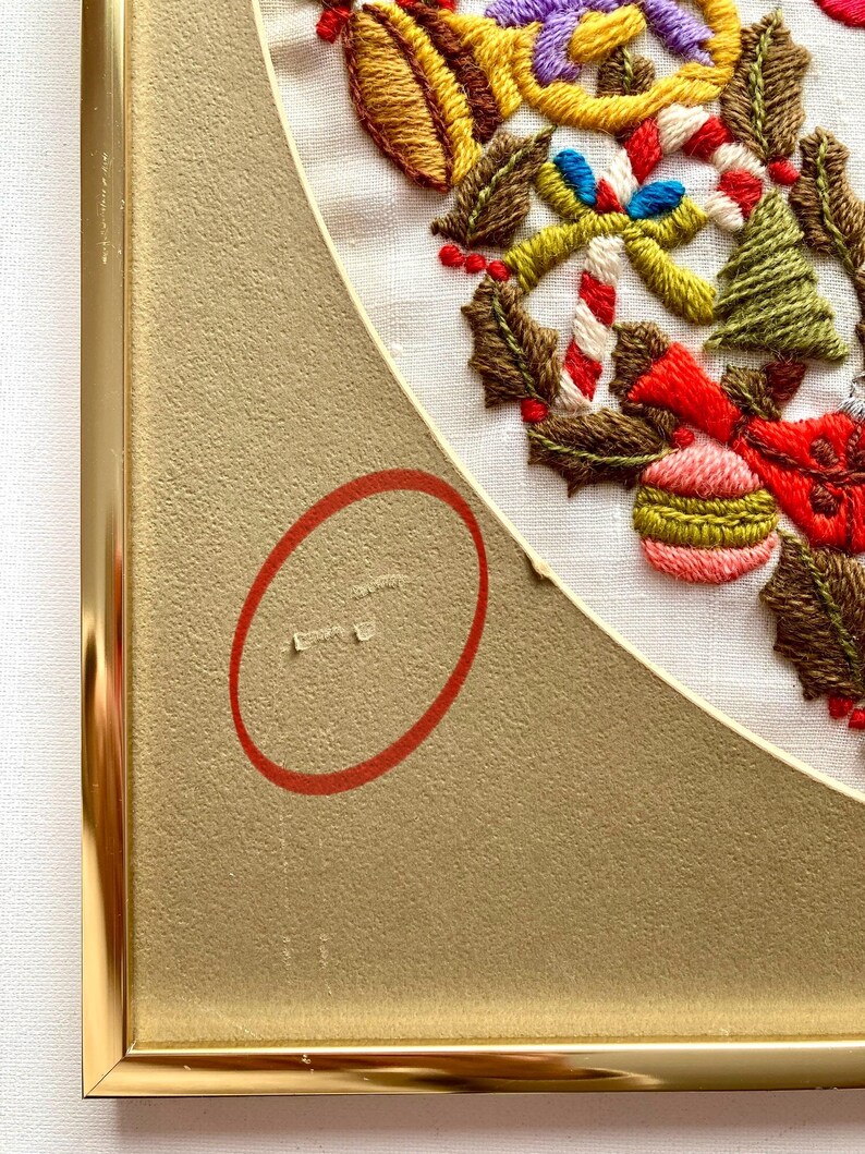 May include: A framed embroidery of a Christmas wreath with holly, candy canes, baubles, and a bell. The wreath is stitched in various colours of thread on white fabric. The frame is gold.
