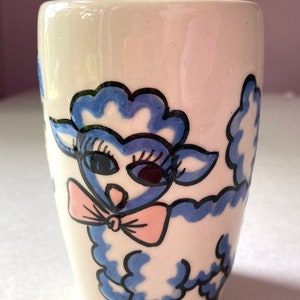 May include: A white ceramic mug with a blue and black illustration of a sheep wearing a pink bow. The sheep has large eyes and a fluffy body.