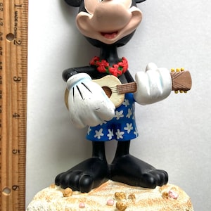 May include: A black and white Mickey Mouse figurine playing a ukulele. He is wearing a blue and white Hawaiian shirt and a red flower lei. He is standing on a white sandy beach with pink and gold accents.