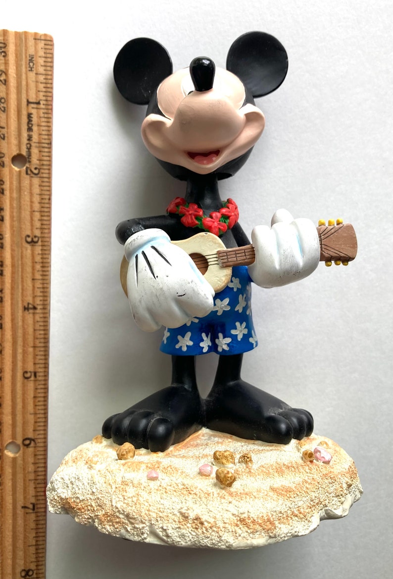 May include: A black and white Mickey Mouse figurine playing a ukulele. He is wearing a blue and white Hawaiian shirt and a red flower lei. He is standing on a white sandy beach with pink and gold accents.