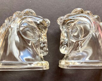 May include: A pair of clear glass horse head bookends. The bookends are facing each other and are made of clear glass. The horse heads are detailed and have a realistic look.