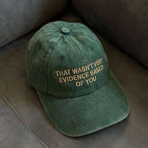 May include: Green baseball cap with the text "THAT WASN'T VERY EVIDENCE BASED OF YOU" embroidered in gold. The cap is made of a soft, textured fabric and has a curved brim. The cap is sitting on a gray surface.