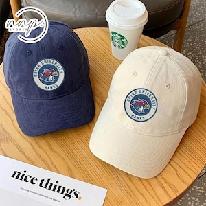 May include: Two baseball caps, one navy blue and one off-white, each featuring a circular patch with "Briar University Hawks" text and a hawk graphic. The caps are on a wooden surface, with a coffee cup and a magazine with the text "nice things."