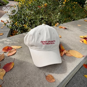 Peut inclure: Une casquette de baseball blanche avec le texte rouge brodé I'M NOT DRAMATIC I'M THEATRICAL sur le panneau avant. La casquette présente une calotte structurée, une visière incurvée et de petits œillets d'aération sur le dessus.