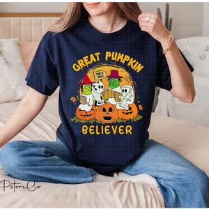 May include: A navy blue t-shirt featuring a Halloween design. The graphic includes cartoon ghosts, a skeleton, and pumpkins, with the words "GREAT PUMPKIN BELIEVER" in yellow. The text "Since 1966" is also present.