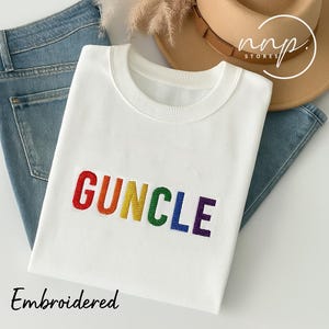 May include: White sweatshirt with the word "GUNCLE" embroidered in rainbow colors. The word is stitched in a rainbow pattern, with each letter a different color. The sweatshirt is folded and laid on a blue denim background.