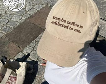 May include: A tan baseball cap with the embroidered phrase "maybe coffee is addicted to me." The hat has a curved brim and a classic six-panel design. The hat is made of a soft material and has a casual style.