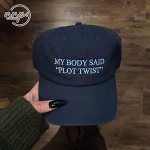 Pode incluir: Um boné de beisebol azul-marinho com texto bordado em azul claro na parte frontal que diz MY BODY SAID PLOT TWIST. O boné possui uma copa estruturada, uma aba curva e ilhós de ventilação na parte superior dos painéis para um visual clássico.