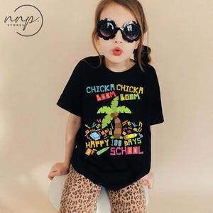 Pode incluir: Uma camiseta preta com o texto "Chicka Chicka Boom Boom" e "Happy 100 Days of School" em letras coloridas. A camiseta apresenta um gráfico de palmeira e outras ilustrações com tema escolar. A criança está usando calças com estampa de leopardo e óculos de sol em forma de flor.