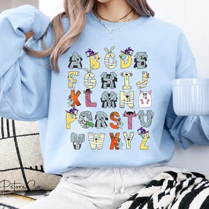 May include: Pale blue sweatshirt with a Halloween-themed alphabet design. Each letter is illustrated with cartoon animals and characters in costumes, in various colours. A casual, comfortable style of sweatshirt.