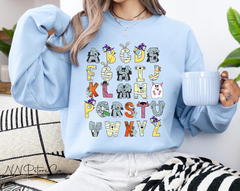 May include: Pale blue sweatshirt with a Halloween-themed alphabet design. Each letter is illustrated with cartoon animals and characters in costumes, in various colours. A casual, comfortable style of sweatshirt.