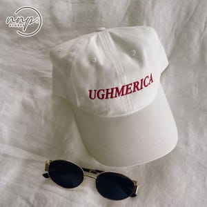 May include: A white baseball cap featuring the red embroidered text UGHMERICA on the front panel, accompanied by a pair of gold-rimmed sunglasses with dark lenses positioned directly below the cap.