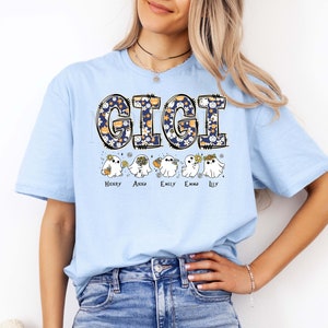 K&ouml;nnte beinhalten: Ein hellblaues T-Shirt mit dem Schriftzug GIGI in einer dunkelblauen Musterschrift mit K&uuml;rbissen und Blumen. Unter dem Text befinden sich f&uuml;nf wei&szlig;e Geisterillustrationen, die jeweils kleine Gegenst&auml;nde halten, mit den Namen Henry, Anna, Emily, Emma und Lily darunter.