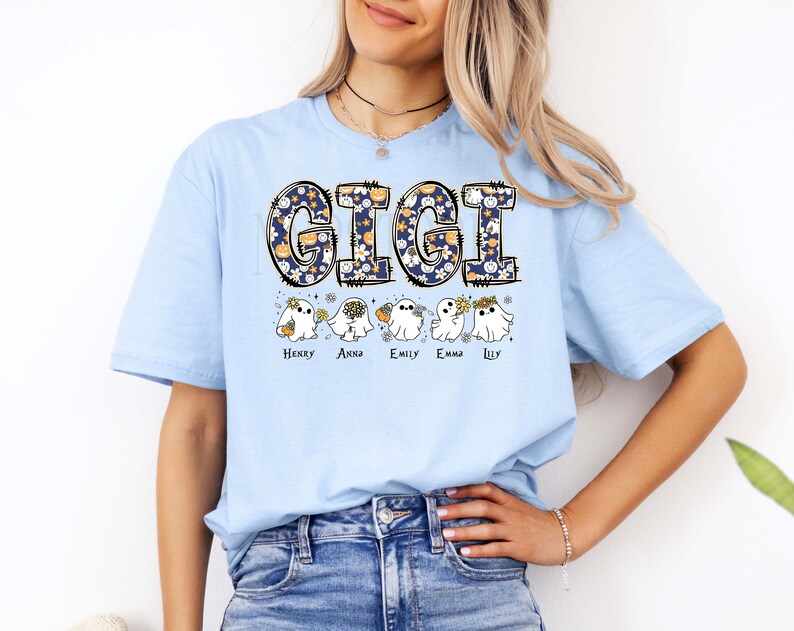 K&ouml;nnte beinhalten: Ein hellblaues T-Shirt mit dem Schriftzug GIGI in einer dunkelblauen Musterschrift mit K&uuml;rbissen und Blumen. Unter dem Text befinden sich f&uuml;nf wei&szlig;e Geisterillustrationen, die jeweils kleine Gegenst&auml;nde halten, mit den Namen Henry, Anna, Emily, Emma und Lily darunter.