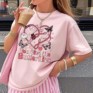 May include: A light pink t-shirt with a graphic design featuring a heart, butterflies, and the text "You Give Me Butterflies" in a playful font. The design includes a lock, key, and other romantic elements.