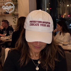 Puede incluir: Una gorra de béisbol blanca con el texto "PROTECT YOUR DAUGHTER" tachado, seguido del texto "EDUCATE YOUR SON" bordado en letras de color rojo oscuro en el panel frontal. La gorra tiene una visera curva y una corona estructurada.