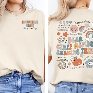 Può includere: Una maglietta beige con una grafica colorata sul retro che dice "Caro membro dello staff che legge questo..." con un arcobaleno, stelle e cuori. La parte anteriore della maglietta ha il testo "Istruttore di formazione Sig.ra Finley" in lettere colorate.