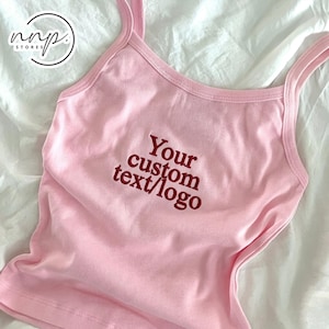 Pode incluir: Uma regata rosa claro com alças finas e decote redondo. O centro da peça apresenta um texto bordado em vermelho escuro que diz Your custom text/logo. O tecido tem uma textura lisa e uma cor pastel sólida.
